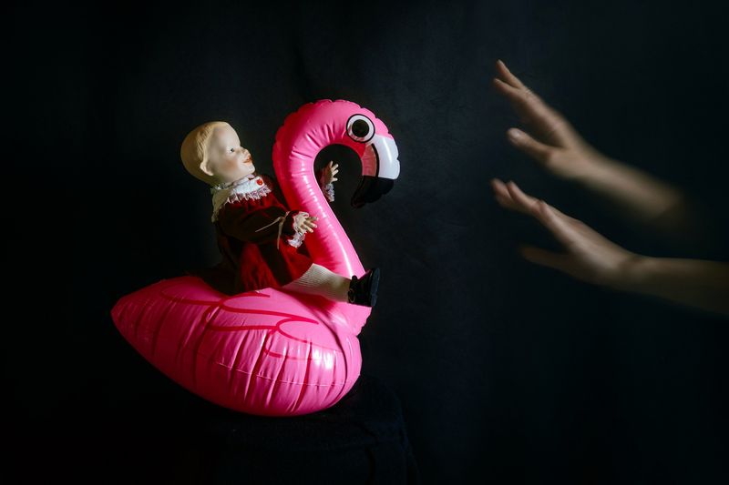 © Mari Saxon - It's too early for you (The doll arrives on an inflatable flamingo, but barely seen hands hold her back, driving her away.)