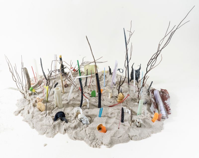 © Florence Iff - arrangement of collected micro‑plastics with organic sea matter found on a beach, embedded in  sand