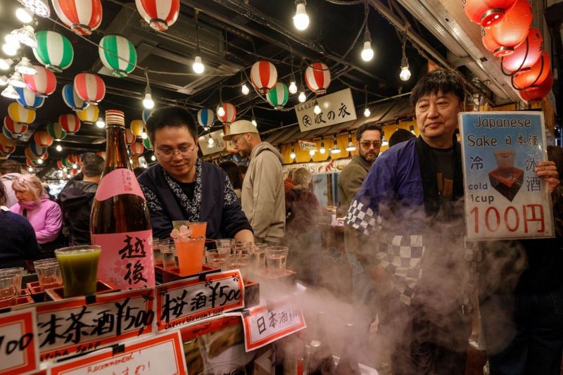 © ROBERTA LODI - "Sake Market Scene"   Vendors offering sake samples in a lively and crowded urban market.