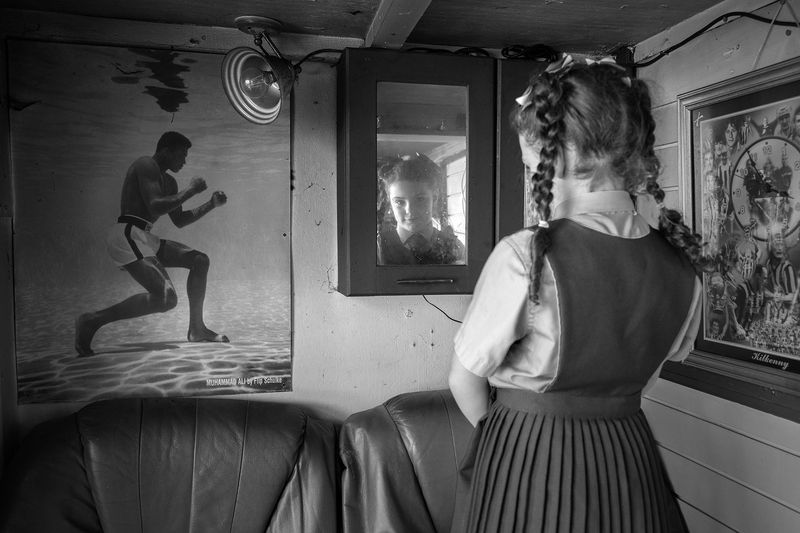 © Rebecca Moseman - A young traveler gazes into a mirror, showcasing her father's shed behind their family caravan.