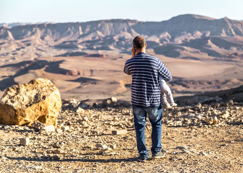 © Nikolay Epifanov - The Phonecall. A man speaking on the phone near a cliff edge with a baby in his arms, the Negev desert, Israel, 2017.