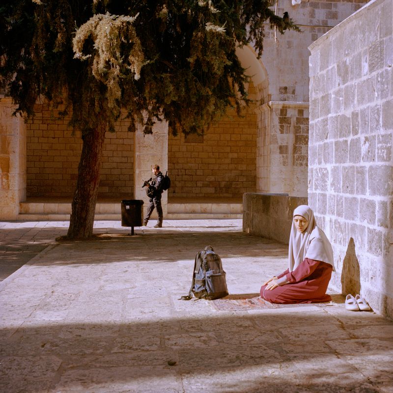 © Fabrizio Bilello - A Palestinian woman praying by the Al-Aqsa mosque, while armed soldiers roam the area. Jerusalem, December 2024.