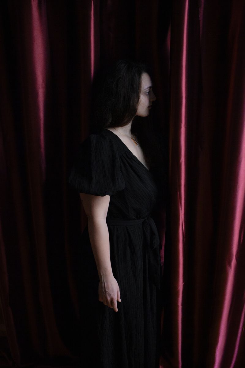 © Susanna Fiona Murray - A solitary figure stands behind a curtain in a dark interior. The image evokes the idea of waiting and transition.