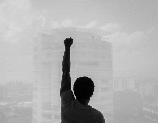 © Roxanne Munson - Black woman with her left fist in the air looking out of a large window.