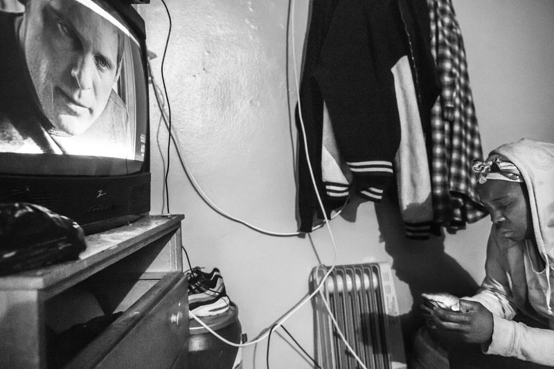 © Nicolas Enriquez - Karen, mother of Kirell and aunt of Smokey looks at her phone wile watching TV inside her apartment in Brooklyn.
