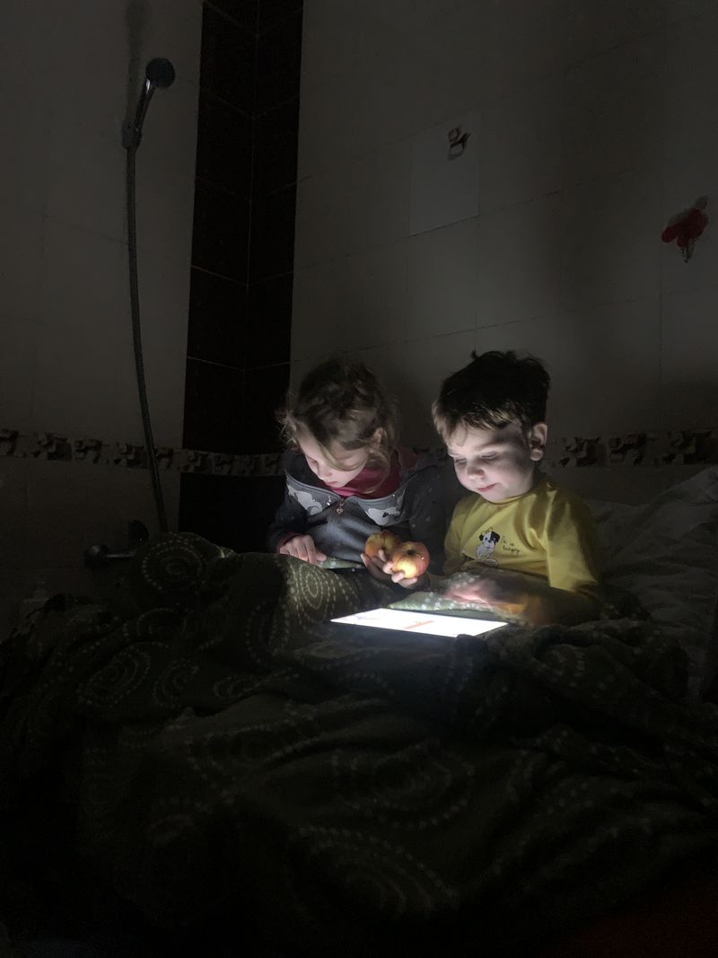 © Ira Sozanska - Tablets in the dark In the bathroom with no light or power, children sit with their tablets, hiding from another attack.