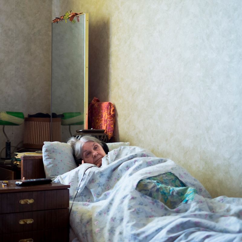 © Maria Quigley - Babushka lays in bed watching her television. On top of her tall mirror are the ribbons of Saint George tied into bows.