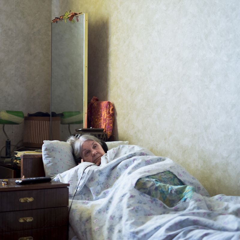 © Maria Quigley - Babushka lays in bed watching her television. On top of her tall mirror are the ribbons of Saint George tied into bows.