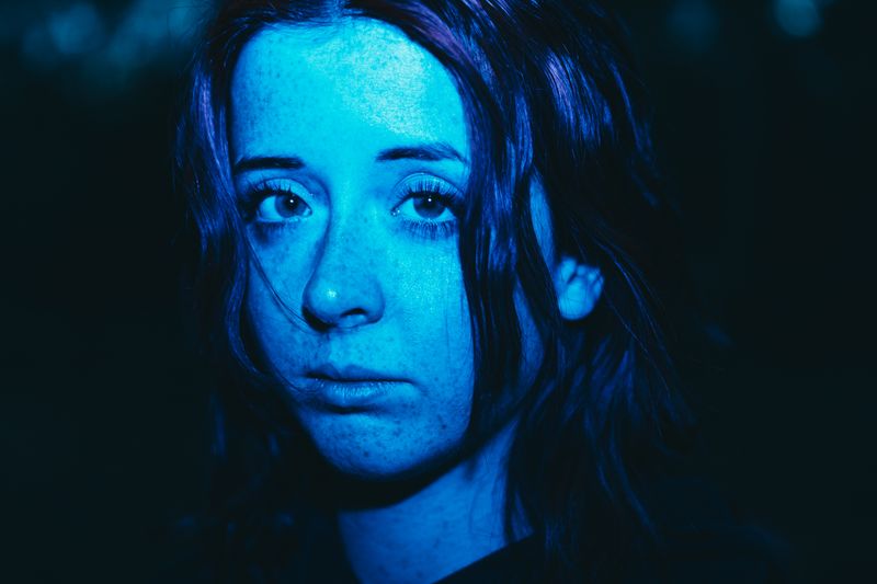 © Ryley Clarke - Close-up portrait of a young woman with blue lighting, dark hair, and freckles.