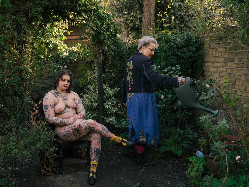 © Fotómetro - Carey watering the plants in the garden while Lois is posing nude to the camera.
