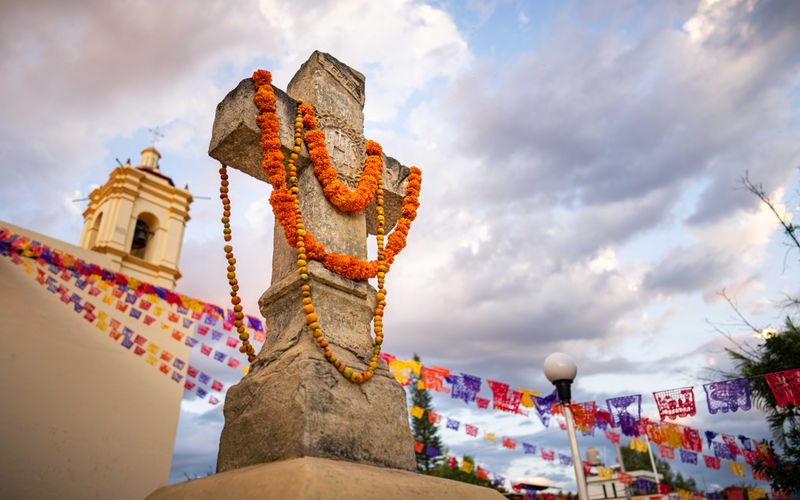 © Hong Yu Chan - A cross wrapped in marigolds stands against the sky, marking the path between worlds.