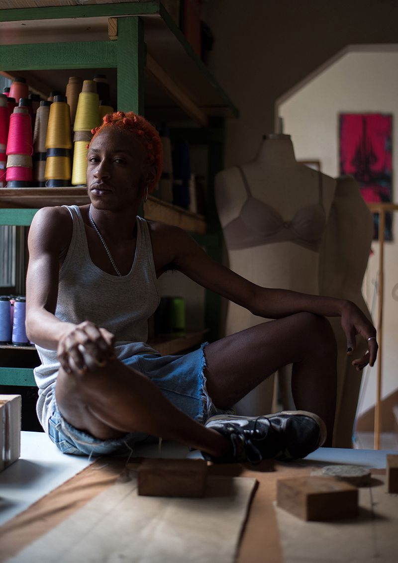 © Camila Falcão - Trindade, waiter and fashion designer, sits on a table in his studio