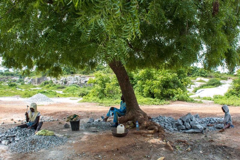 © Igho Ogbobine - Using the tree as shade, children sit to break rocks.
