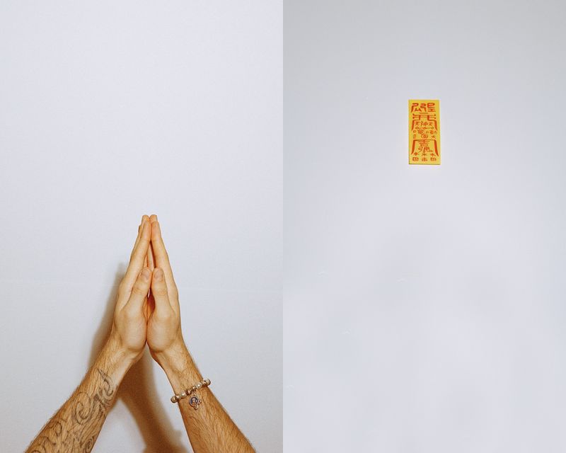 © Juno Joo - Hands gathered in prayer beside a protective charm, two systems of belief meet in quiet coexistence.