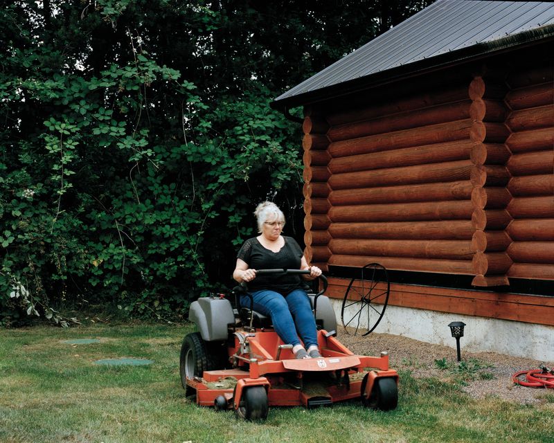 © Marco Waldis - A woman is mowing the lawn in front of a log cabin that she and her husband built to rent out as a bnb.