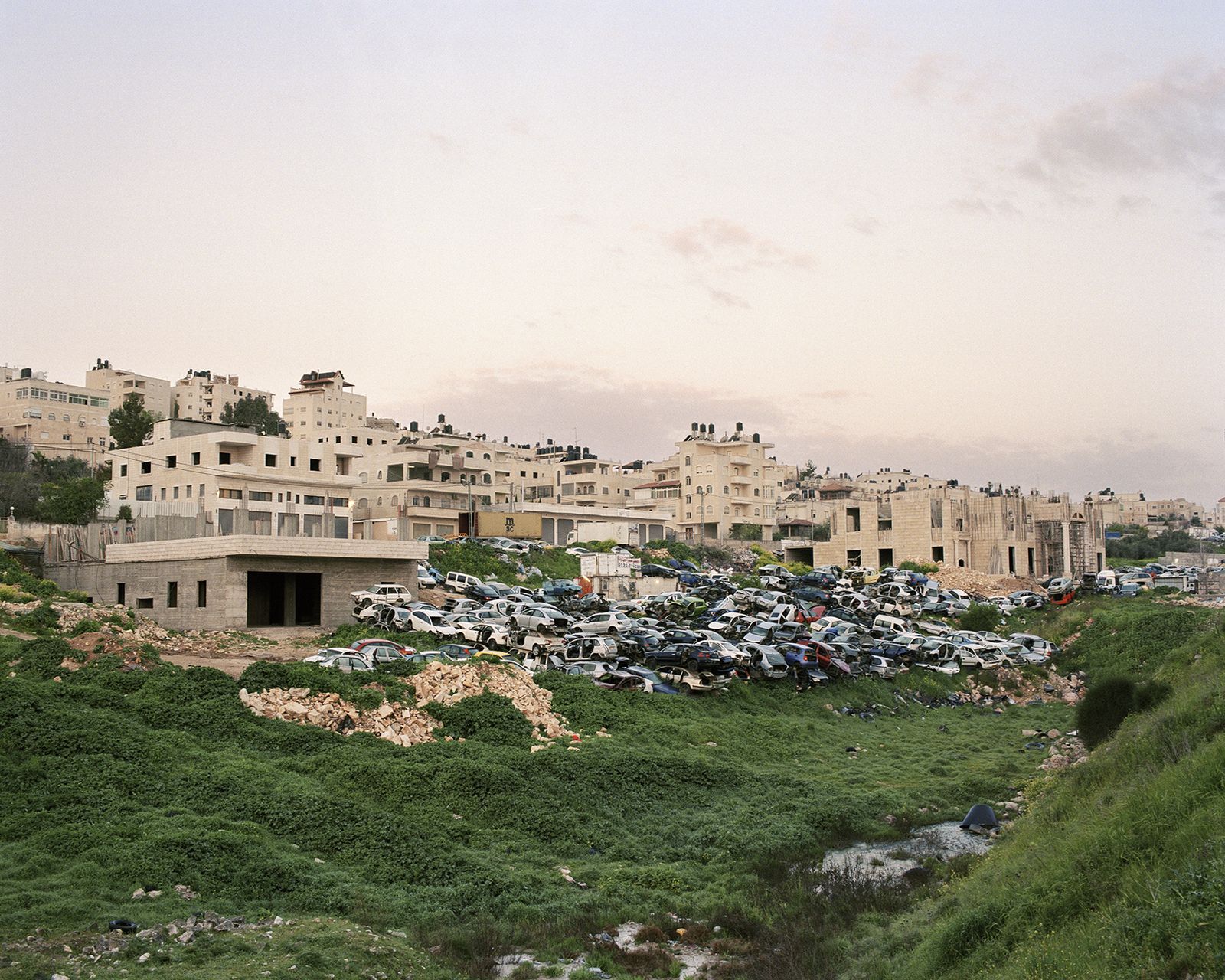 © Saja Quttaineh - A mountain of neglected cars (Car graveyard) and behind it a disorganized building in Al-Ram town, 2021.