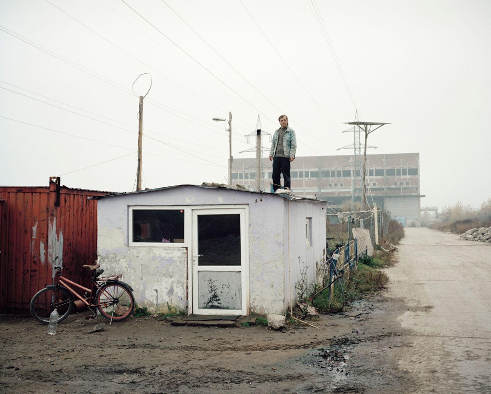 © Tommaso Rada - Romania, Giurgiu. A man working as security guard stand on a barrack in front of an abandoned factory that he watch.