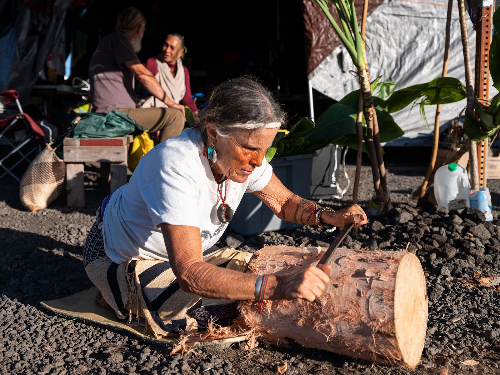 © Molly Peters - Aunty Clare, a midwife, works on carving a drum near the Kūpuna tent. Aunty Scarlett and Uncle Jon chat in the background.