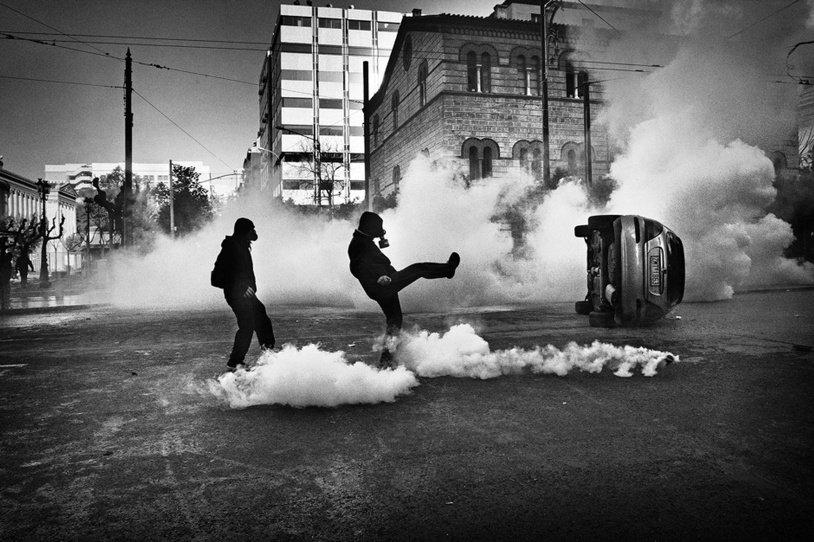 © Nikos Pilos - 2008. Two protesters wearing gas masks are kicking away a tear-gas canister.