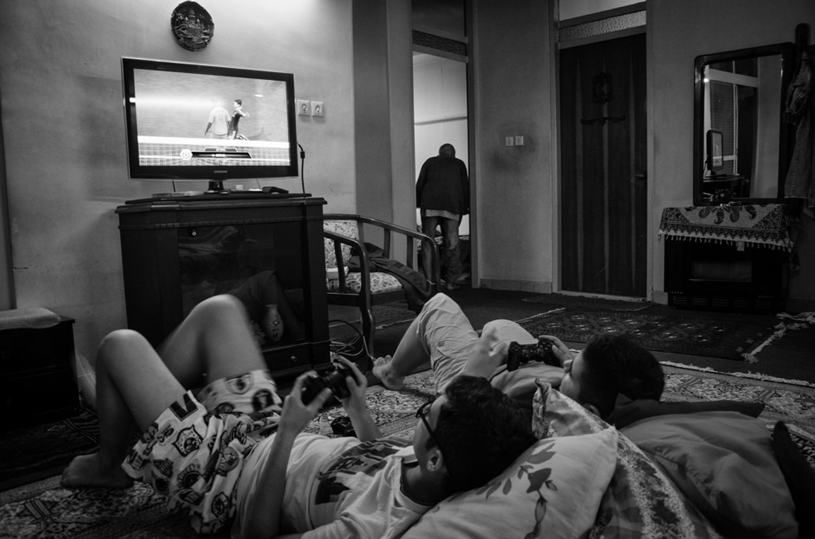 © Khashayar Sharifaee - Grandchildren are playing with Playstation, grandfather is trying hardly to put on his pants after coming out of bathroom.